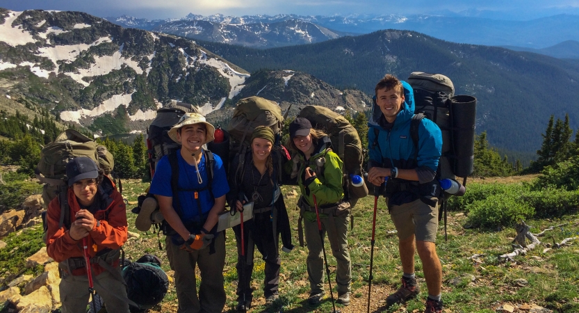 Five students with backpacks smile with a mountainous landscape in the background